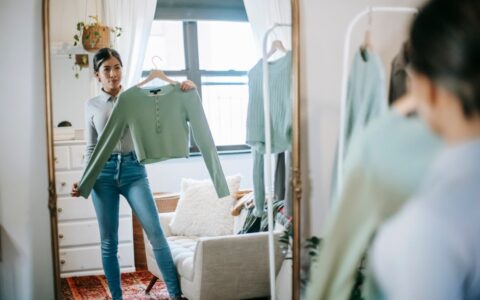 Serious young ethnic woman in trendy outfit choosing and demonstrating blouse on hanger in room near couch and cabinet at light home near wardrobe and looking in mirror