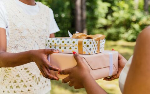 Two individuals exchanging colorfully wrapped gift boxes in a lush outdoor setting, symbolizing celebration.