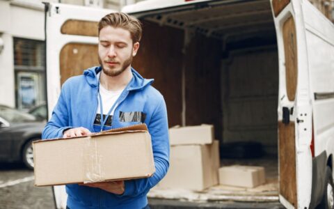 A young delivery man carrying a package near an open delivery truck in the city street.
